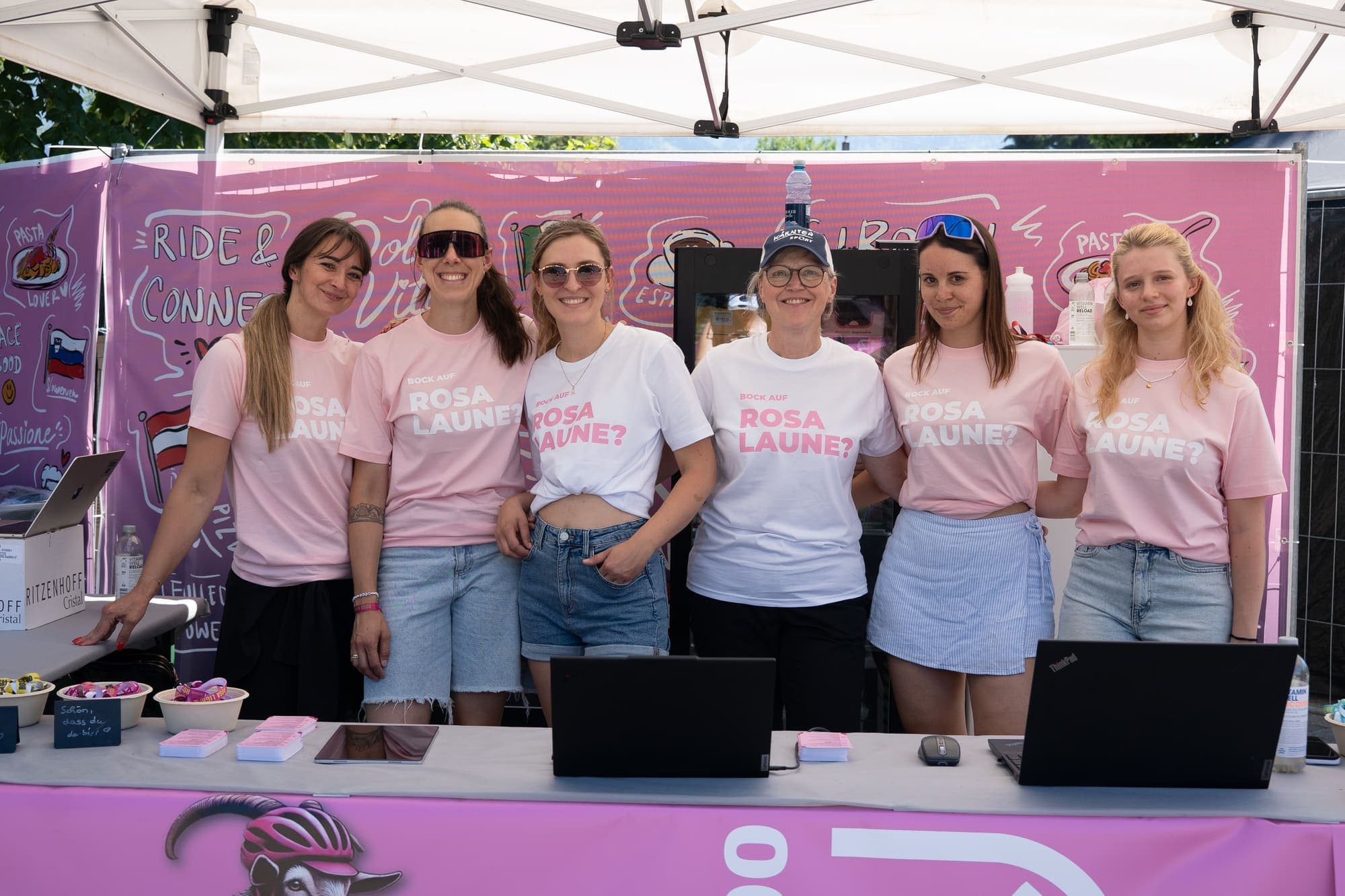 Sechs Helferinnen in rosa Shirts mit der Aufschrift Rosa Laune hinter einem Stand mit Laptops